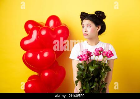 Bonne Saint Valentin.Triste et solitaire fille regardant gauche bouleversée, tenant bouquet de roses, debout seul près des ballons rouges de coeur Banque D'Images