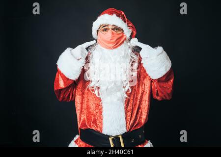 Homme habillé comme le Père Noël avec un masque rouge, pointant vers lui avec ses doigts, sur fond noir.Concept de Noël, Père Noël, cadeaux, celebbratio Banque D'Images