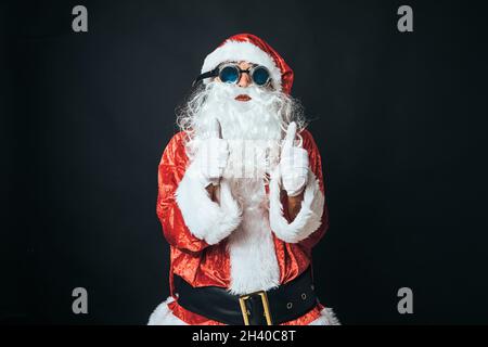 Homme habillé comme le Père Noël avec des lunettes de soudeur de style victorien, levant les pouces, sur fond noir.Concept de Noël, Père Noël, cadeaux, cele Banque D'Images