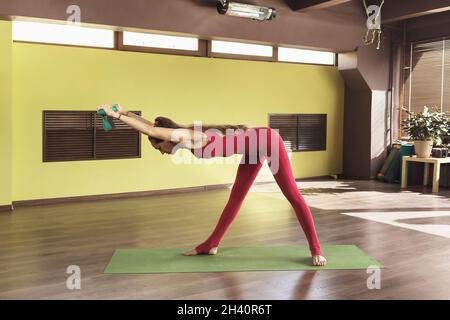 Une femme effectue un exercice trikonasana avec des lanières, posture de triangle, se tient seul sur un tapis dans le studio dans une combinaison rouge de sport Banque D'Images