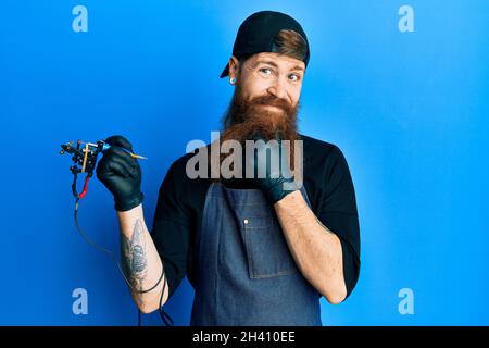 REDHEAD homme avec la longue barbe tatouage artiste portant l'uniforme professionnel et les gants regardant avec confiance la caméra souriant avec les bras croisés et la main r Banque D'Images