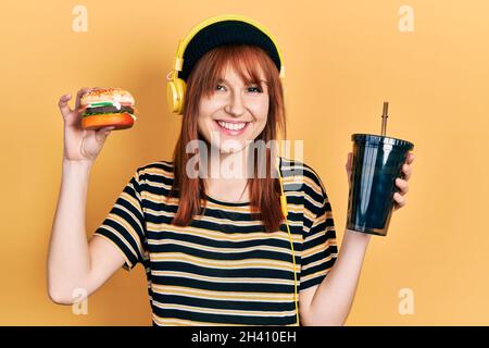 REDHEAD jeune femme mangeant un hamburger et buvant du soda portant des écouteurs souriant avec un sourire heureux et frais sur le visage. Montrant des dents. Banque D'Images