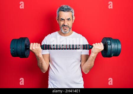 Beau homme d'âge moyen avec des cheveux gris portant des vêtements de sport en utilisant des haltères expression sans cluess et confuse. Doute concept. Banque D'Images