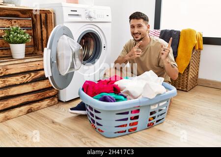 Jeune homme charmant mettant du linge sale dans le lave-linge souriant et regardant la caméra pointant avec deux mains et les doigts sur le côté. Banque D'Images