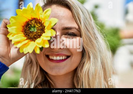 Jeune femme blonde souriant avec du tournesol à l'œil au parc Banque D'Images