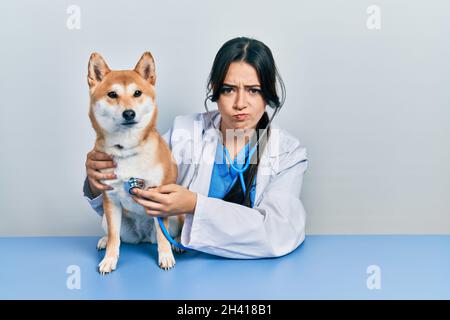 Belle femme hispanique vétérinaire vérifiant la santé de chien sceptique et nerveux, frowning bouleversé à cause du problème. Personne négative. Banque D'Images