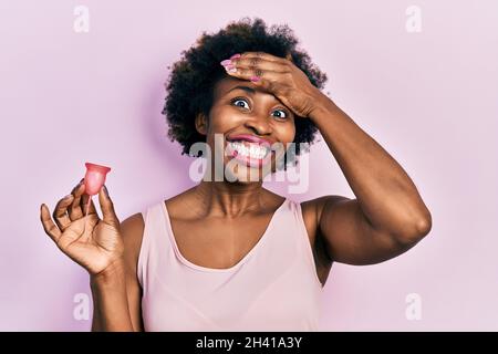 Jeune afro-américaine tenant une coupe menstruelle stressée et frustrée avec la main sur la tête, surprise et colère visage Banque D'Images