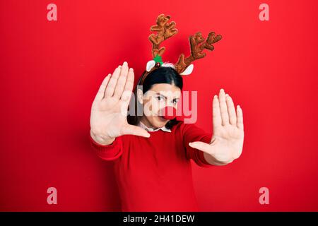 Jeune femme hispanique portant un chapeau de noël de cerf et nez rouge faisant le cadre avec les mains paumes et les doigts, la perspective de caméra Banque D'Images