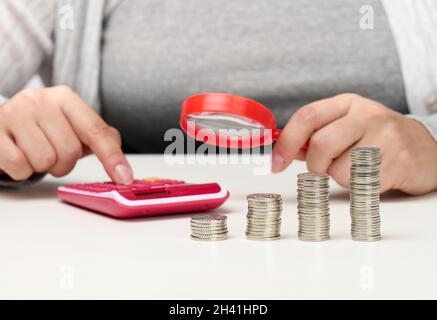 Une femme s'assoit à une table et compte sur une calculatrice de l'autre main une loupe, une pile de pièces de monnaie sur la table.Calcul Banque D'Images