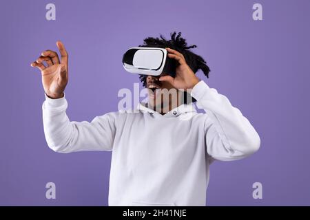 Un jeune homme afro-américain émerveillé dans un casque VR utilisant la réalité virtuelle, touchant l'écran imaginaire sur fond violet Banque D'Images