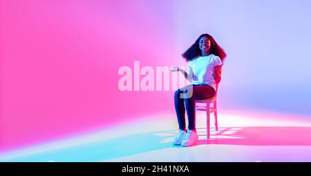 Bonne dame noire assise sur la chaise et offrant un espace vide pour votre annonce dans la lumière de néon, le design de bannière Banque D'Images