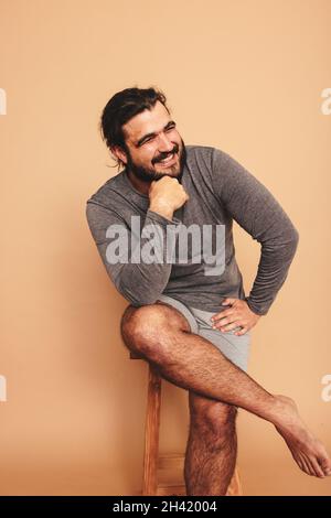 Joyeux jeune homme souriant gaiement dans un studio.Corps positif jeune homme regardant loin tout en étant assis sur une chaise en bois.Sentiment de jeune homme d'auto-assurance c Banque D'Images