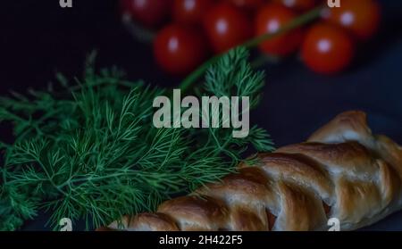 Saucisses roulées de saucisses cuites dans une pâte feuilletée, tomates sur fond sombre. Banque D'Images