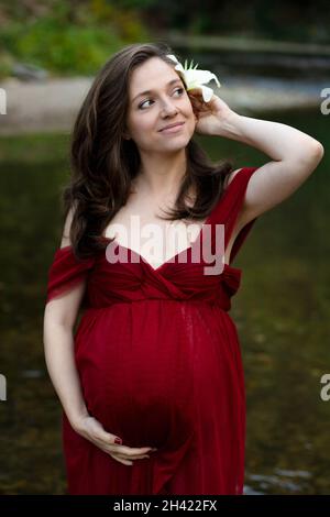 Jeune femme enceinte blonde, élégante, debout dans la rivière en robe rouge, fleur blanche derrière son oreille Banque D'Images