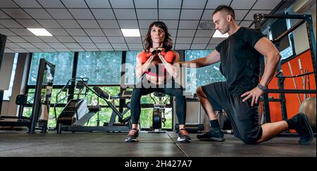 Belle femme athlétique avec kettlebell faisant une fente.Sport fille fait des exercices tout en s'exerçant avec un entraîneur personnel dans la salle de gym - force an Banque D'Images