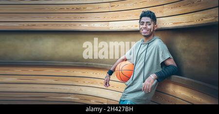 Adolescent américain positif avec la coiffure afro il regarde et souriant à l'appareil photo - Portrait de beau garçon noir tenant le ballon de basket-ball en plein air Banque D'Images