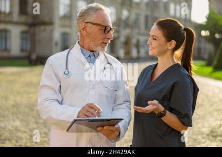 Portrait d'un médecin de laboratoire en blouse et en lunettes tenant le presse-papiers, en regardant une jeune infirmière souriante, debout ensemble à l'extérieur près de la clinique Banque D'Images