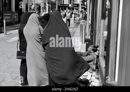 Cardiff, pays de Galles le 31 2021 juillet : une jeune femme sans domicile assise sur le trottoir du centre-ville de Cardiff.Un groupe de femmes asiatiques dans les hijabs s'arrêtent de parler Banque D'Images