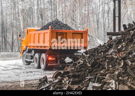 Un camion à benne basculante dans une zone industrielle ou sur un chantier de construction en hiver décharge le charbon cokéfiant de la benne, à côté d'une pile de déchets métalliques et de déchets. Banque D'Images