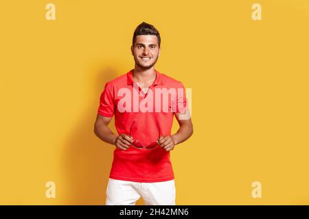 Un jeune homme taille basse en t-shirt rouge et short blanc tient des lunettes de soleil tandis que se tient sur fond jaune Banque D'Images