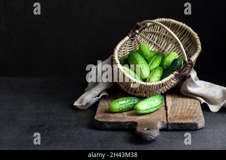 Concombres frais dans un panier en osier sur panneau en bois sur fond noir. Banque D'Images