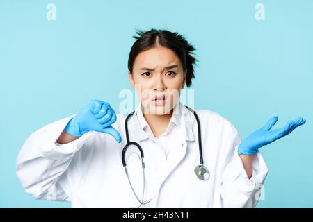 Frustré asiatique médecin, l'infirmière montre les pouces vers le bas et regarde confus, ne comprend pas le patient, debout dans l'uniforme médical sur fond bleu Banque D'Images