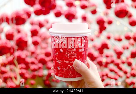 Main de femme tenant une tasse à café à thème de vacances avec des ornements boule rouge floue en arrière-plan Banque D'Images