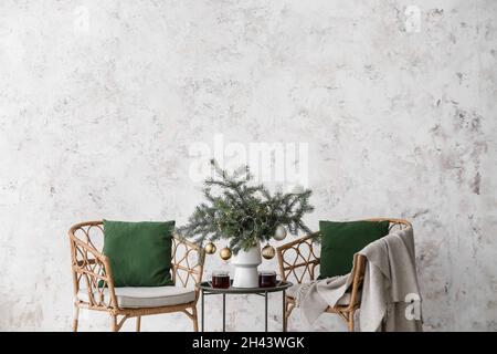 Branches de sapin avec boules de Noël dans un vase, tasses de thé sur une table et chaises près du mur lumineux Banque D'Images