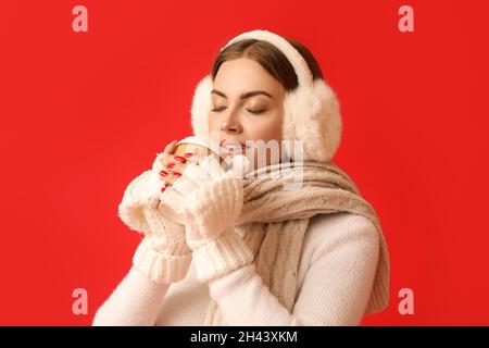 Belle jeune femme en casque chaud buvant du café sur fond rouge Banque D'Images