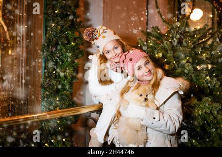 Deux jeunes filles sourient et s'amusent à tenir le chien spitz en regardant la décoration de la rue de noël.Belles sœurs, arbre de noël décoré et lumières sur fond.Une soirée magique dans la rue Banque D'Images