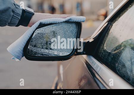 Main de l'homme lave le rétroviseur de la voiture avec un chiffon en microfibre, essuyant les gouttes d'eau Banque D'Images