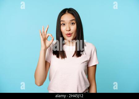 Une jeune fille coréenne brune enthousiaste montre un signe correct, regardant smth fantastique, prix génial, recommander un magasin, debout sur fond bleu Banque D'Images