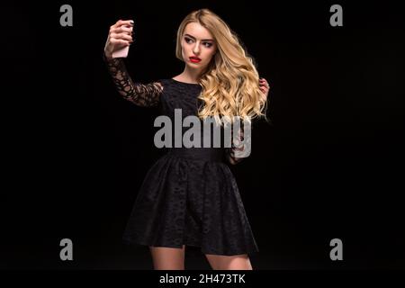 Belle femme blonde en robe noire et maquillage prendre selfie avec téléphone mobile isolé sur le fond noir Banque D'Images