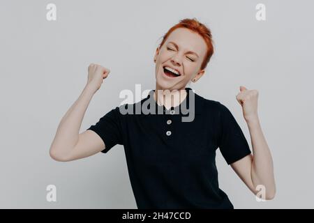 Une femme aux cheveux rouges surjoyeuse garde les yeux fermés et crie de l'excitation contre le mur gris Banque D'Images