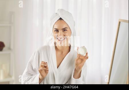 Portrait de femme souriante et mignonne dans un peignoir et une serviette tenant la crème du visage dans sa main. Banque D'Images