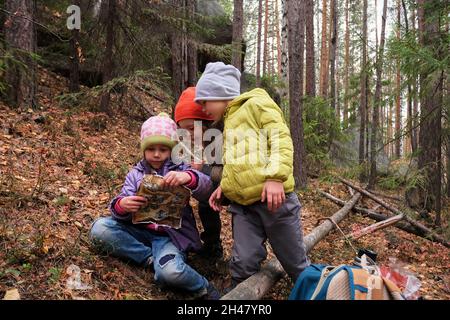 Groupe d'enfants en vêtements d'extérieur chauds examinant la carte tout en étant assis dans des bois sur des feuilles mortes pendant le camping dans la nature d'automne Banque D'Images