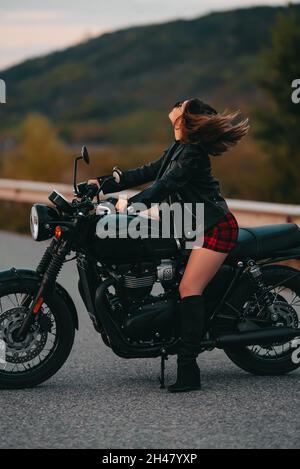 Portrait d'une femme moto assurée en mini jupe, veste en cuir sur une moto de style rétro.Un conducteur attrayant dans des bottes de protection sur route Banque D'Images