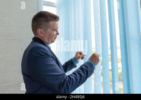 Un jeune homme regardant à travers les aveugles. Un homme d'affaires en costume bleu regarde par la fenêtre à travers les stores Banque D'Images