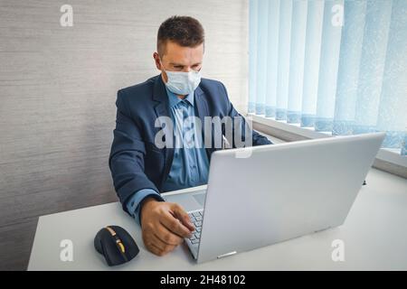 un jeune homme, portant un masque médical et une veste bleue, est assis sur une chaise et travaille sur un ordinateur portable dans une pièce lumineuse. bureau internet Banque D'Images