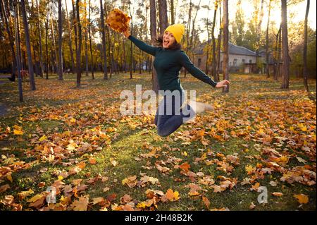 Femme gaie à pull-over vert chaud, laine jaune et chapeau denim décontracté sautant haut avec un bouquet de belles feuilles d'érable d'automne sèches dans les mains, Banque D'Images