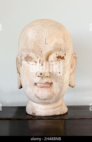 Gros plan sur une tête de Bouddha en bois ancienne ou ancienne avec une peinture de craquage et une expression spirituelle sereine affichée sur une étagère Banque D'Images