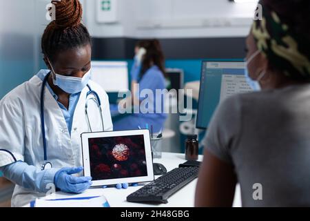 Médecin afro-américain tenant un ordinateur de tablette discutant d'une infection virale expliquant le traitement médical qui fonctionne dans le bureau de l'hôpital.Femmes portant un masque de protection contre la covid19 Banque D'Images