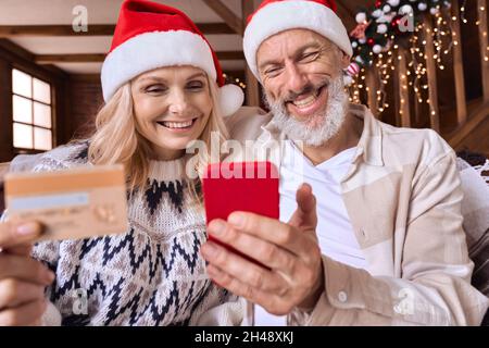 Joyeux vieux couple porter des chapeaux de père noël tenir téléphone carte de crédit faire des achats de Noël. Banque D'Images