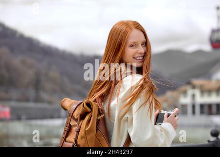 Jeune femme redhead souriant heureux en utilisant vintage caméra marche à la montagne, la femme a tourné la tête en arrière et regarder l'appareil photo, dans une tenue décontractée. Portrai Banque D'Images