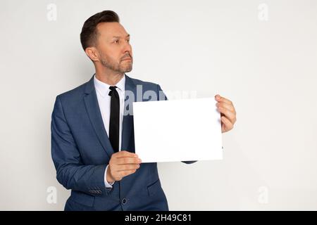 Jeune homme beau en bussines costume avec barbe tenant le papier vide à la main sur fond blanc, regardant à côté.Prise de vue en studio Banque D'Images
