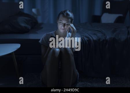 Femme épuisée assise à côté de son lit et vérifiant son smartphone tard la nuit, elle souffre d'insomnie Banque D'Images