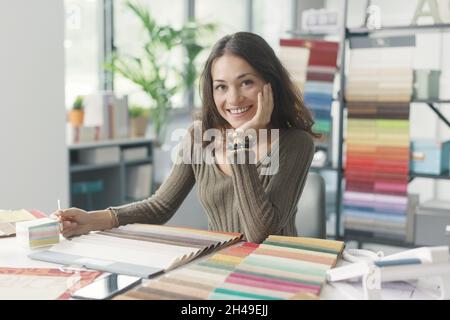 Jeune décoratrice d'intérieur assise au bureau et travaillant dans son studio, elle sourit à la caméra Banque D'Images