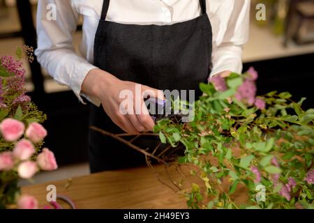 une femelle écourtée tenant des ciseaux tout en coupant des fleurs, préparant un bouquet dans un fleuriste. une femme méconnaissable en tablier noir organise des fleurs sauvages Banque D'Images