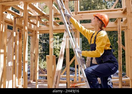 Idées de rénovation et d'extension de maisons en bois.Femelle sur échelle, monte jusqu'au toit.Femme caucasienne travailleur en uniforme de travail et sécurité har Banque D'Images