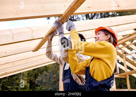 Deux entrepreneurs excités travaillant avec un perceuse visseuse.Menuisier travaillant sur la section de toit de cadre de squelette de maison en bois.Thème de l'industrie de la construction.Femme Banque D'Images